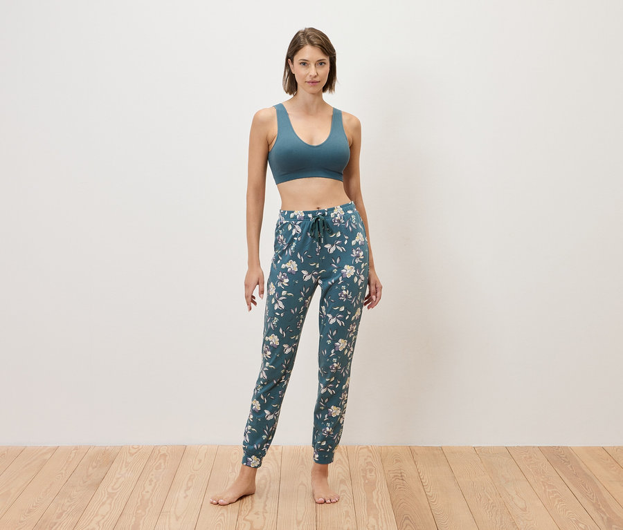 Kadın çıplak ayakla bir duvarın önünde duruyor, mavi bir spor sütyeni ve çiçek desenli mavi eşofman altı giyiyor.