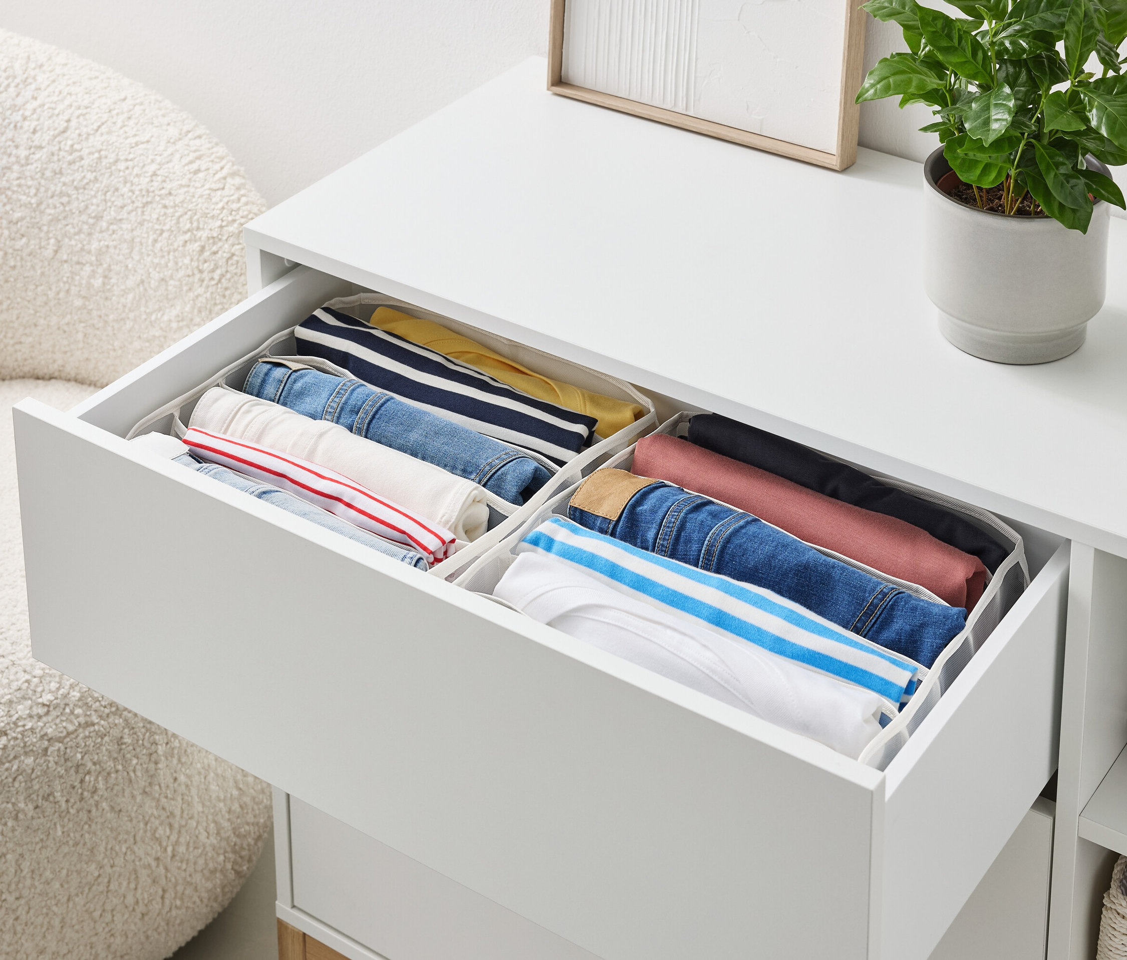 Beyaz bir komodinin açık çekmecesi, düzenleyiciler içinde düzgünce katlanmış giysiler içerir.