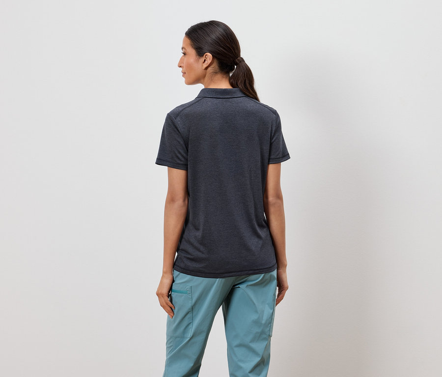 Atkuyruklu bir kadın kameraya arkası dönük duruyor, koyu gri bir polo tişört ve açık mavi pantolon giyiyor.