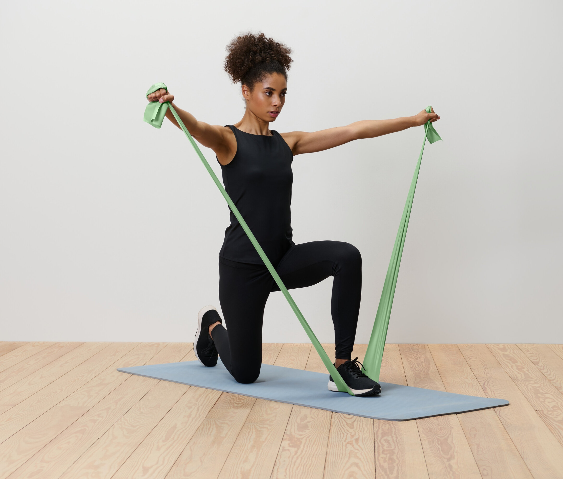 Bir kadın mavi bir yoga matı üzerinde diz çökmüş, yeşil bir direnç bandını geriyor.