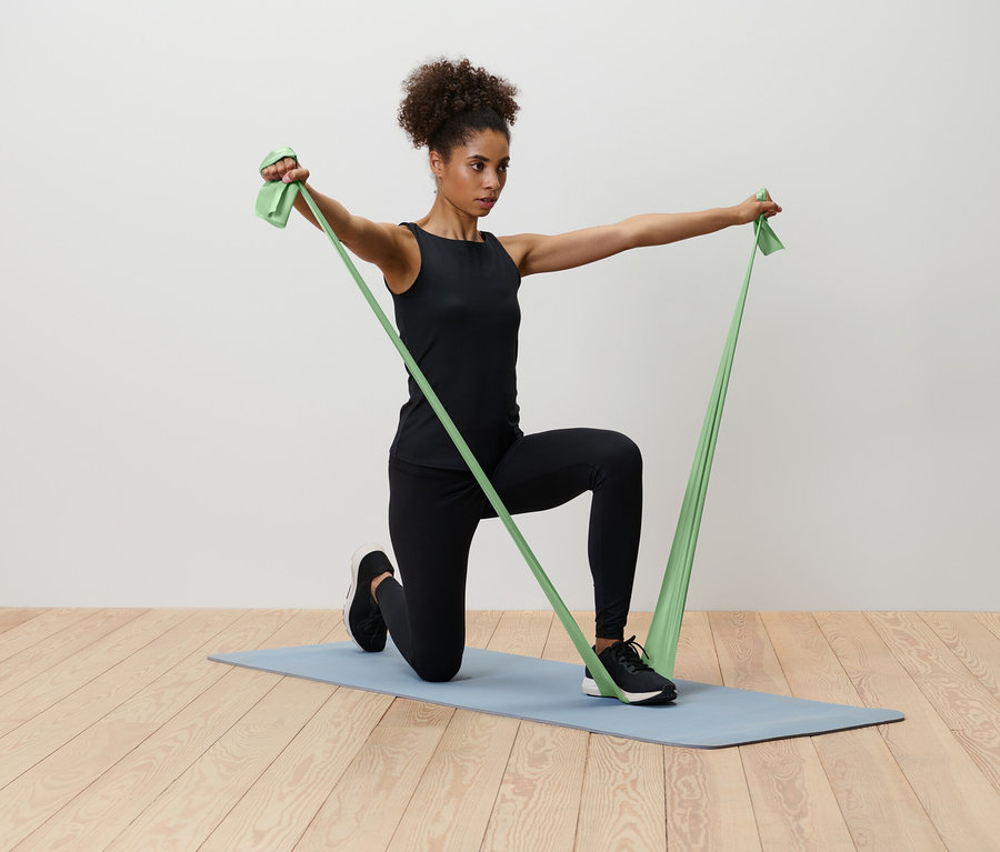 Bir kadın mavi bir yoga matı üzerinde diz çökmüş, yeşil bir direnç bandını geriyor.