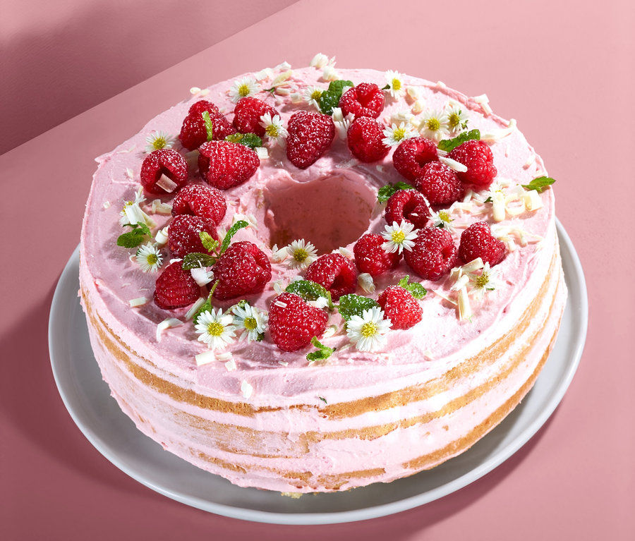 Beyaz tabakta pembe kremalı, ahududulu, çiçekli ve talaşlı çok katmanlı kek.