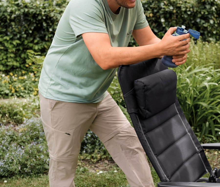 Nane yeşili outdoor tişört ve bej 2'si 1 arada outdoor fonksiyonel pantolon giyen gülümseyen adam, siyah bir bahçe sandalyesine yaslanmış mavi bir su şişesi tutuyor.