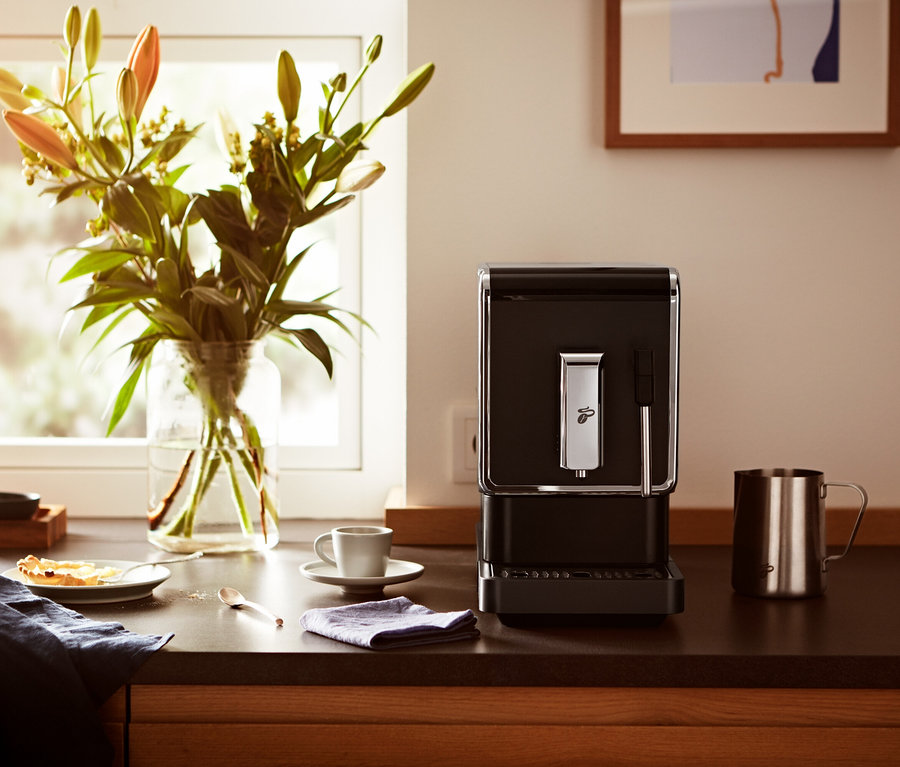 Mutfak tezgahında bir fincan ve çiçek vazosunun yanında antrasit rengi »Esperto Latte« Tchibo Tam Otomatik Kahve Makinesi bulunmaktadır.