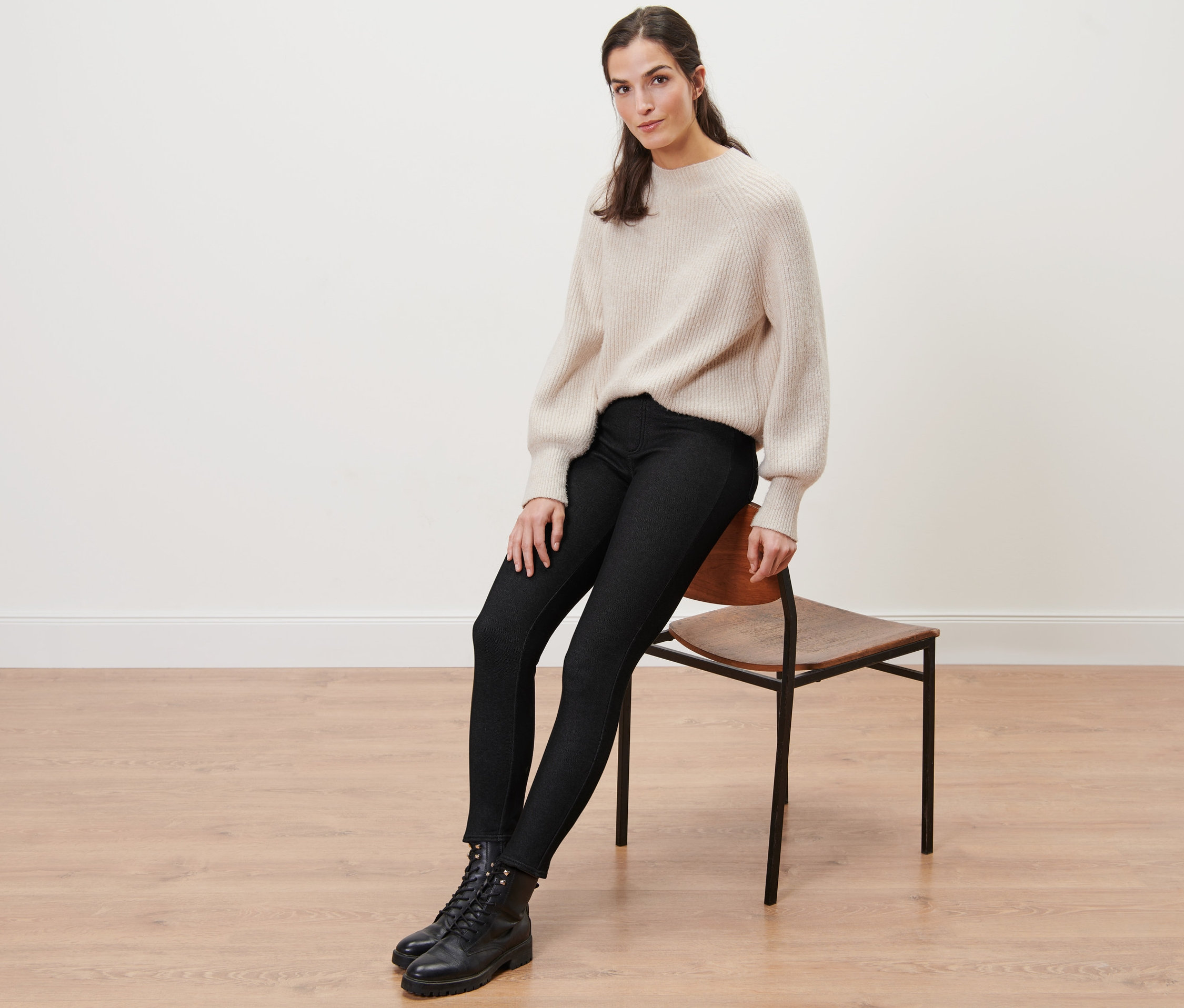 Bir kadın sandalyede oturuyor ve Jean Görünümlü Şardonlu Tayt, kazak ve bot giyiyor.