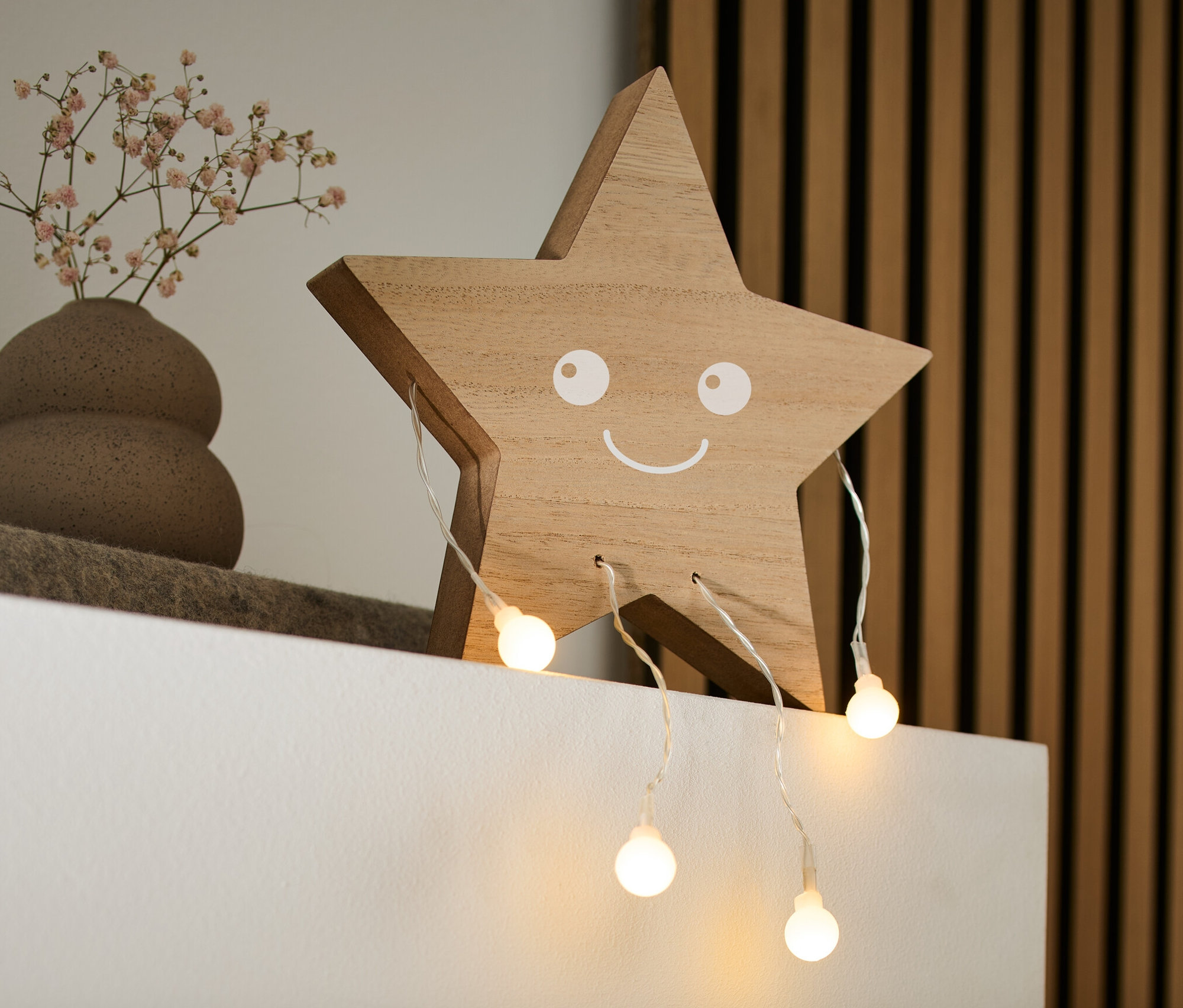 Beyaz bir rafta gülümseyen yüzü olan ahşap bir LED'li Dekoratif Figür »Yıldız«, küçük çiçekli bir vazo ve bir kitap duruyor.
