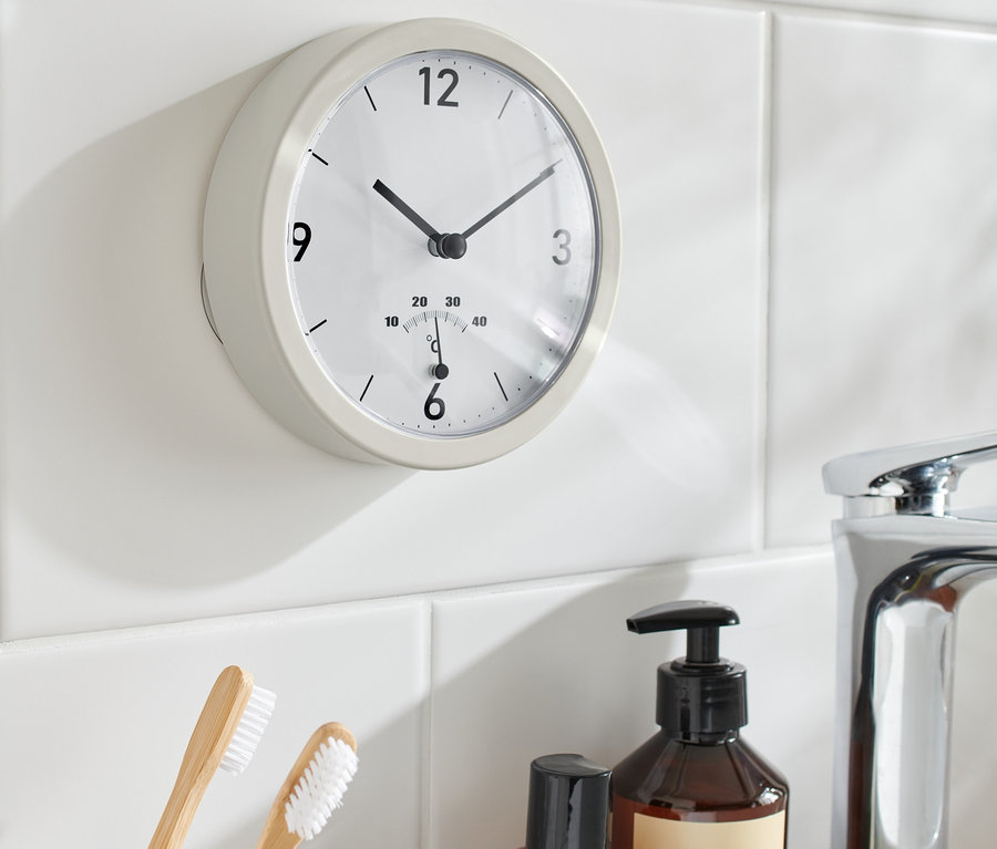 Termometreli banyo duvar saati, lavabo, diş fırçaları ve şişelerin yanında beyaz fayanslara asılı duruyor.