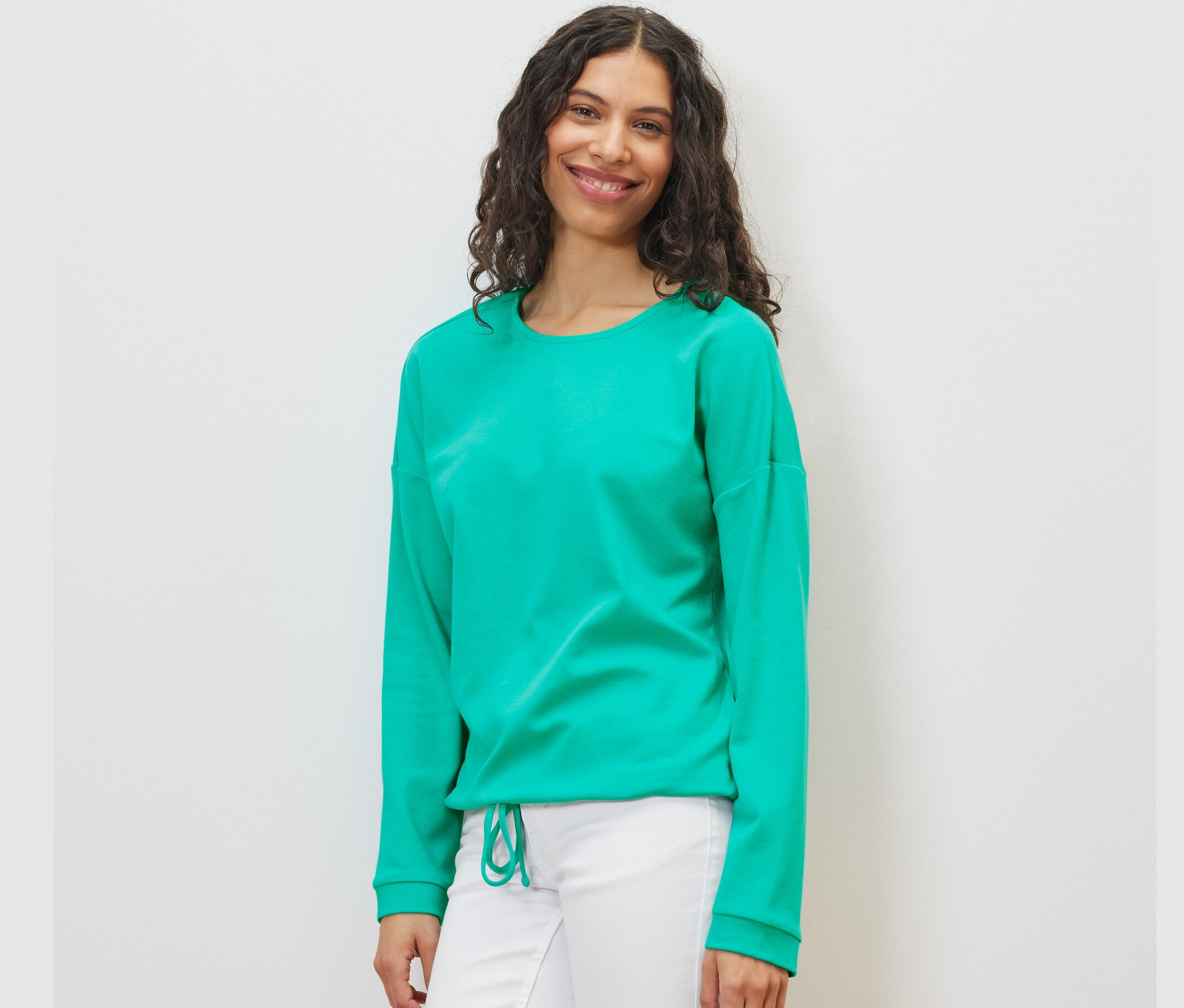 Kadın aqua rengi bir sweatshirt ve beyaz pantolon giyiyor.