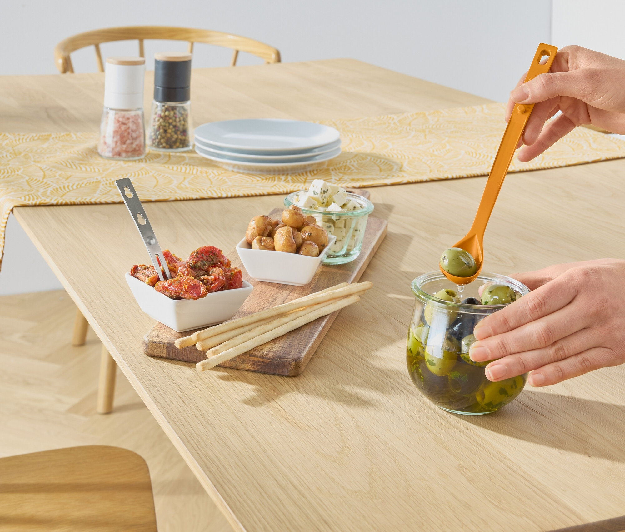 Bir el, tahta bir masanın üzerinde turuncu bir kaşıkla bir kavanozdan yeşil zeytin alıyor. Masanın üzerinde domates, mantar, peynir, çubuklar, tuz ve karabiber değirmenleri bulunan kaseler var.