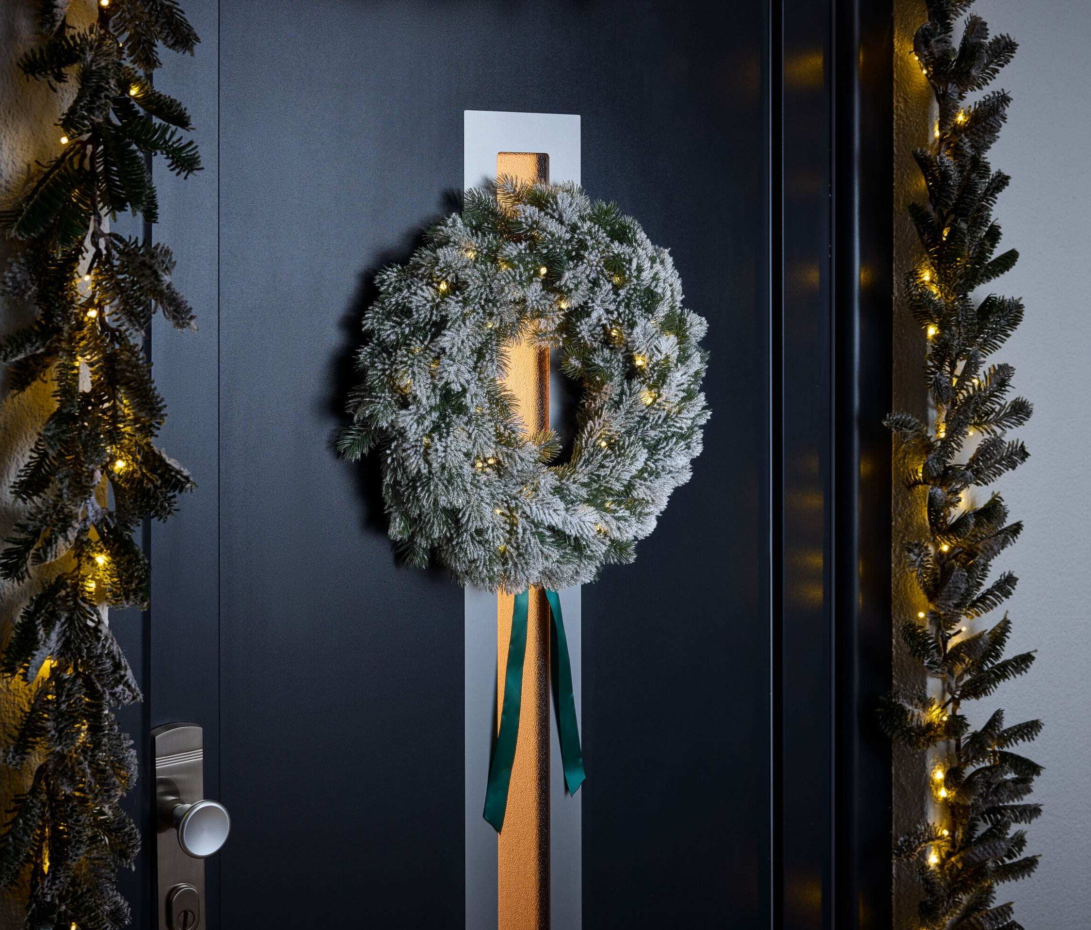 Beyaz toz serpilmiş, yeşil kurdeleli bir çelenk, koyu mavi bir kapıda asılı duruyor. Kapının etrafını küçük ışıklarla süslenmiş çelenkler çevreliyor.