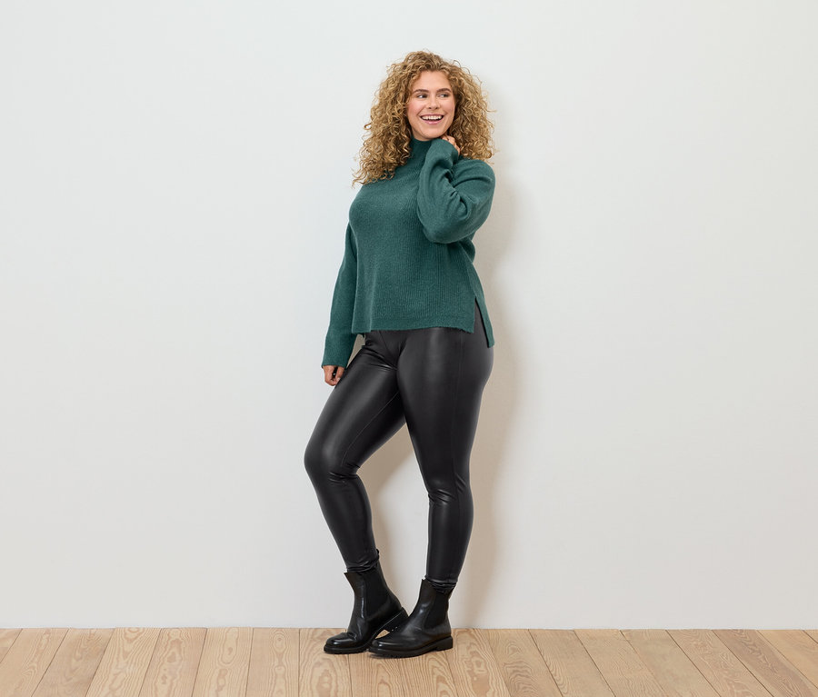 Kıvırcık saçlı kadın, yeşil balıkçı yaka kazak, siyah deri görünümlü tayt ve siyah botlarla poz veriyor.