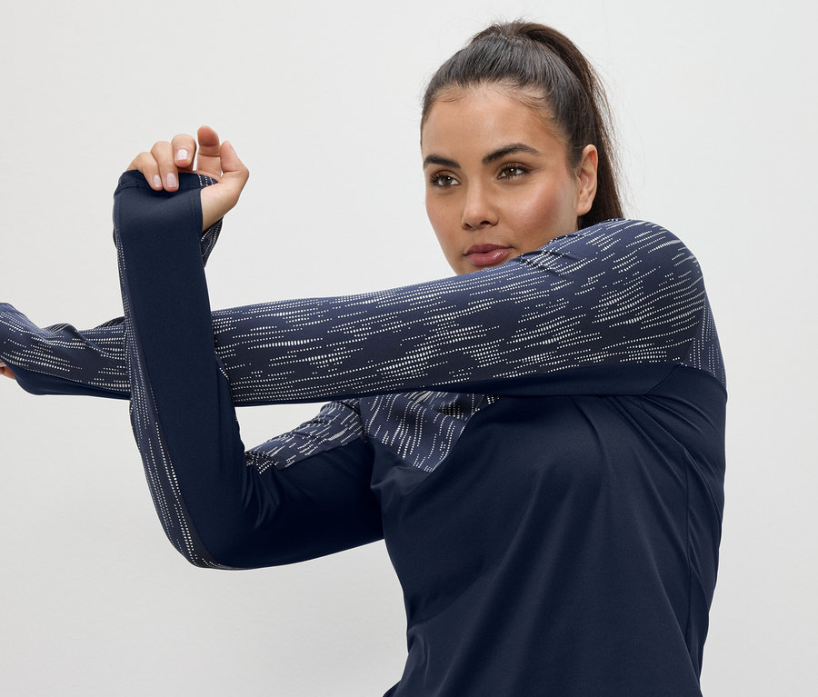 Kadın, lacivert, uzun kollu, başparmak delikli bir spor üstü içinde kollarını geriyor.