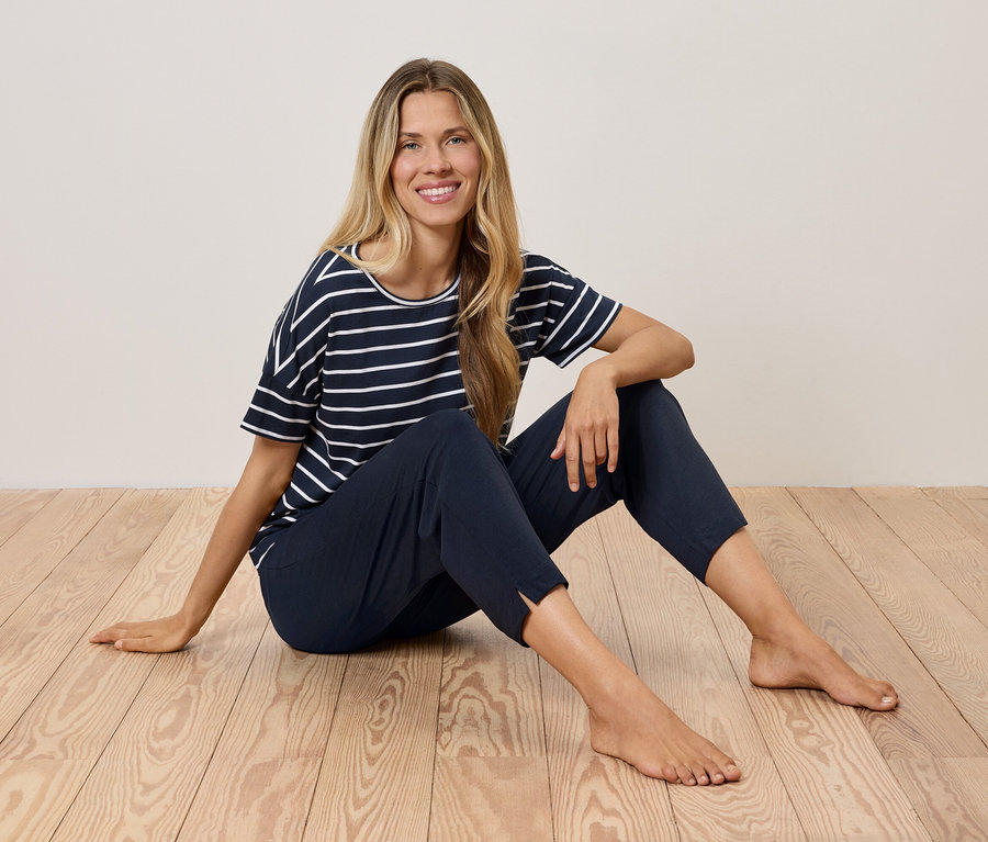 Sarışın bir kadın, ahşap bir zeminde oturuyor, lacivert çizgili bir tişört ve koyu mavi pantolon giyiyor.