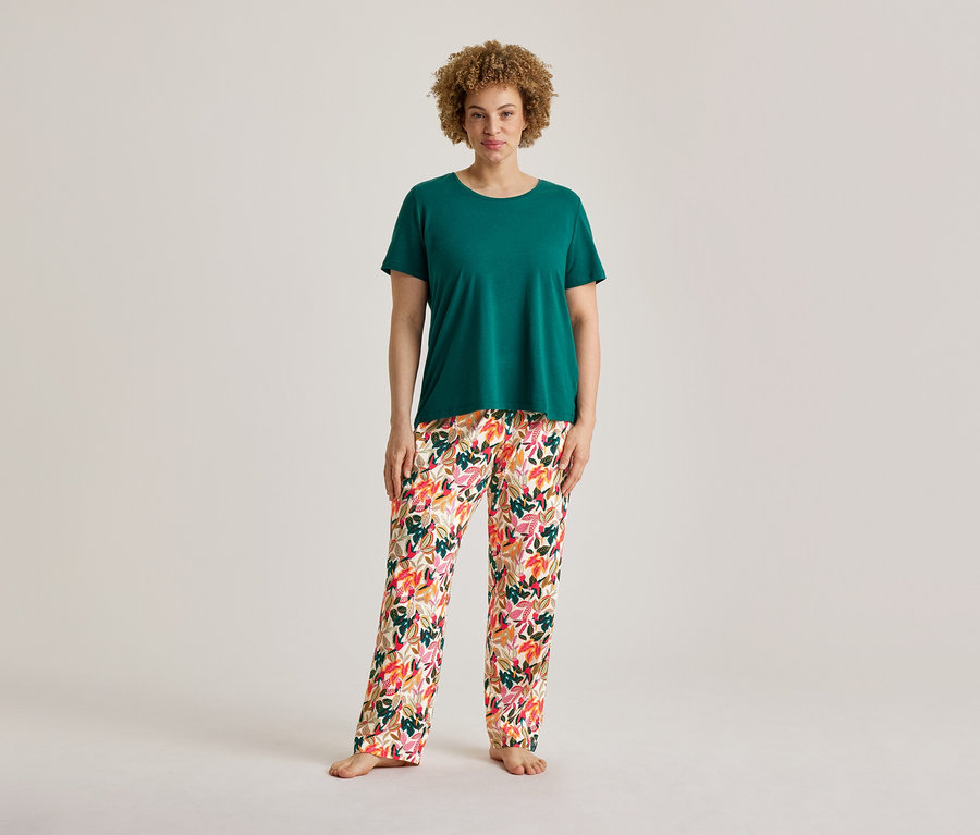 Kıvırcık saçlı bir kadın ayakta duruyor, yeşil bir tişört ve pijama takımından çiçek desenli pijama altı giyiyor, ayakları çıplak.