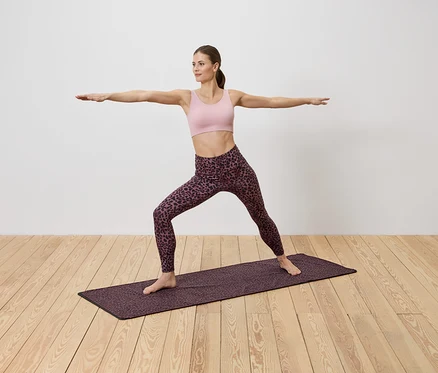 Spor kıyafetli kadın, beyaz duvar önünde ahşap zeminde desenli bir mat üzerinde yoga yapıyor.