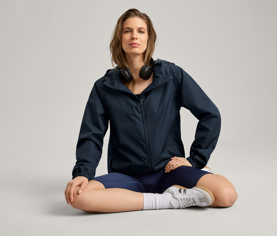 Kadın bağdaş kurmuş oturuyor, üzerinde yağmurluk, kulaklık, şort ve spor ayakkabı var.