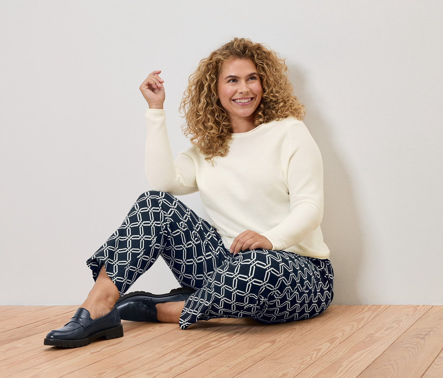 Kıvırcık saçlı bir kadın ahşap zeminde oturuyor, krem rengi bir kazak ve geometrik desenli mavi bir pantolon giyiyor.