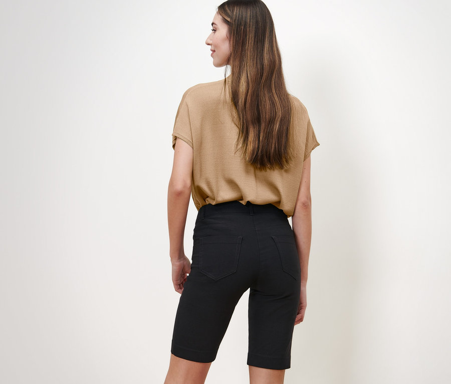 Kameraya arkası dönük duran bir kadın bej bir bluz ve siyah Bengalin Bermuda Şort giyiyor.