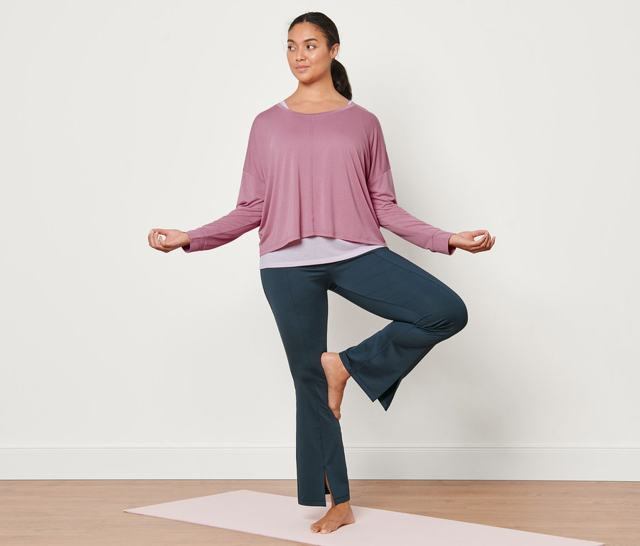 Yoga matında ağaç pozisyonunda duran kadın, koyu mavi bodyforming spor pantolon ve pembe 2'si 1 arada spor tişörtü giyiyor.