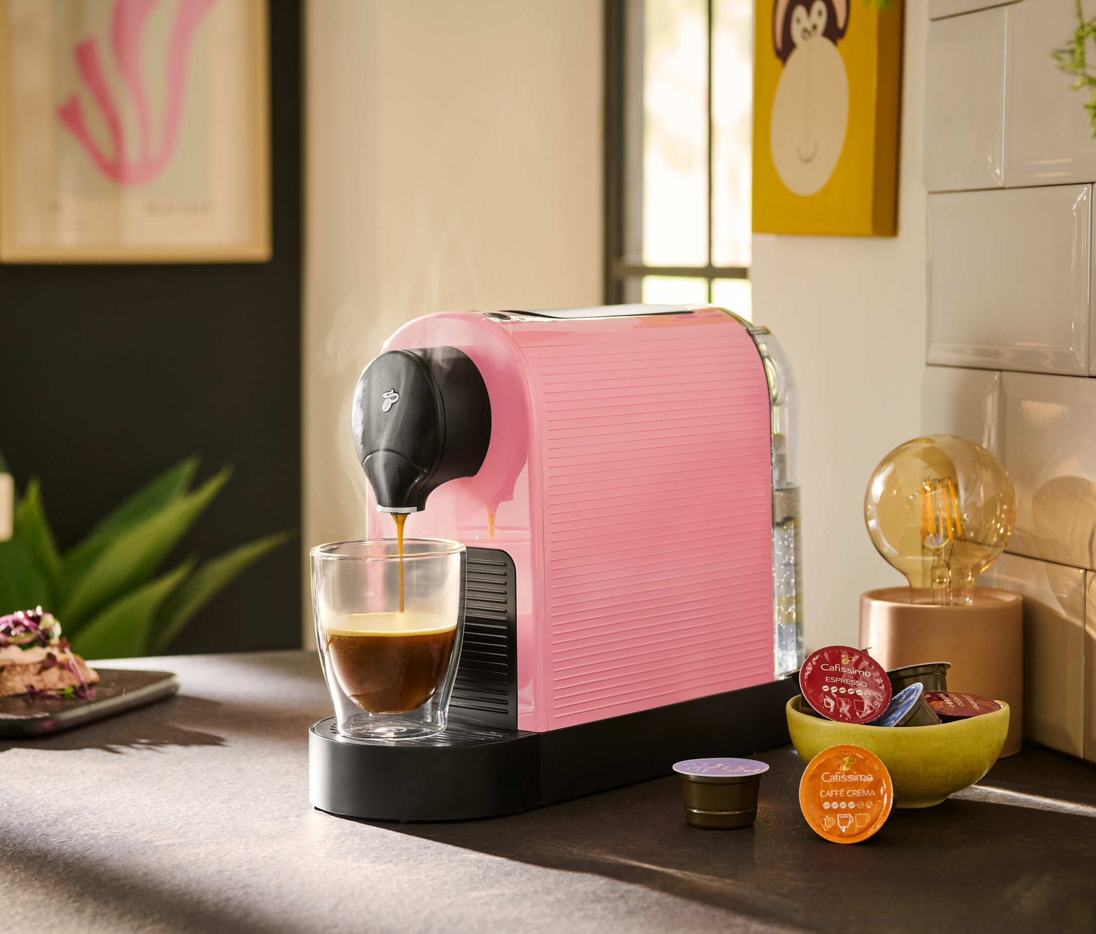 Kiraz çiçeği rengindeki Cafissimo pure+ kahve makinesi bir bardağa kahve koyuyor. Yanında bir kasede kapsüller bulunuyor.