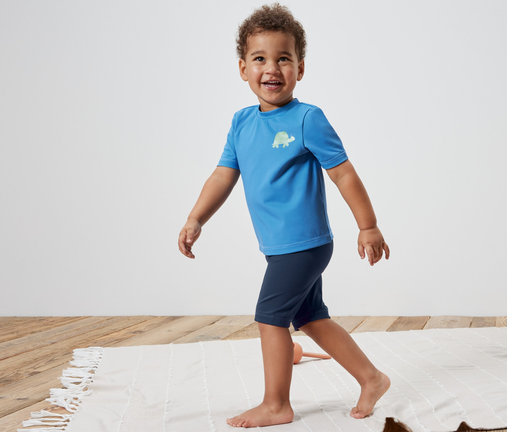 Bir çocuk sahilde 80 Faktör UV Korumalı Çocuk Deniz Taytı ile yürüyor.