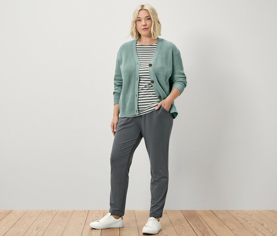 Bir kadın ayakta duruyor ve joggpant, adaçayı renginde kalın örgü bir hırka ve yeşil çizgili, yarım kollu bir tişört giyiyor.