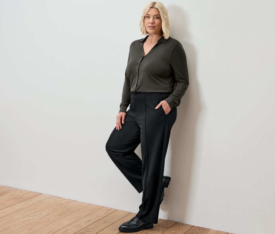 Duvara yaslanmış sarışın bir kadın, siyah dikişli jarse pantolon ve düğmeli bluz giyiyor.