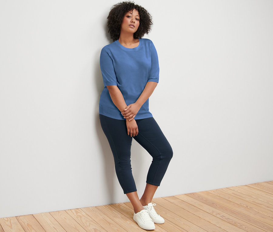 Duvara yaslanmış bir kadın mavi ince triko kazak, lacivert pantolon ve beyaz spor ayakkabı giyiyor.