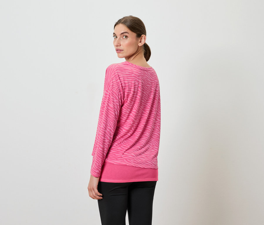 Bir kadın omzunun üzerinden bakıyor, pembe uzun kollu bir üst ve siyah pantolon giyiyor.