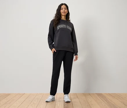 Kadın, elleri siyah eşofmanının cebinde duruyor. Üzerinde Sports Club yazılı gri bir sweatshirt ve beyaz spor ayakkabılar var.