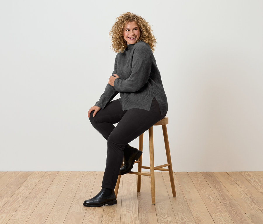 Kıvırcık saçlı bir kadın ahşap bir taburede oturuyor. Gri balıkçı yaka kazak, siyah pantolon ve siyah botlar giyiyor.