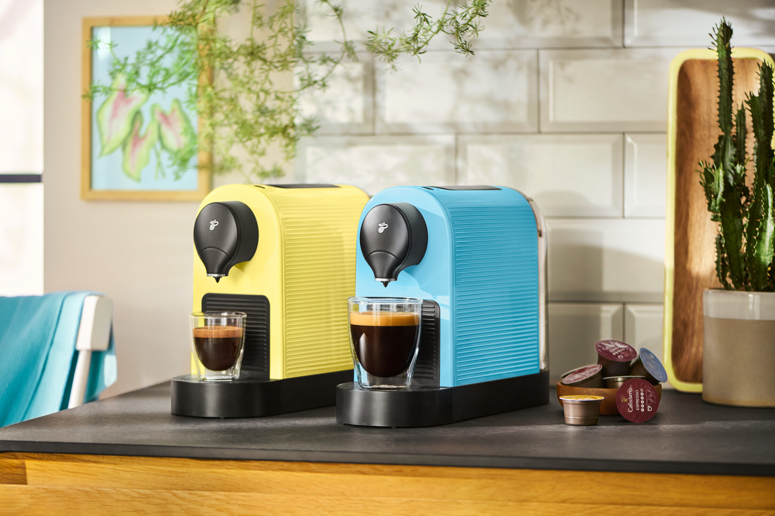 Sarı ve mavi renkte iki kahve makinesi, iki fincan kahve ve kahve kapsülleriyle birlikte bir mutfak tezgahı üzerinde duruyor.