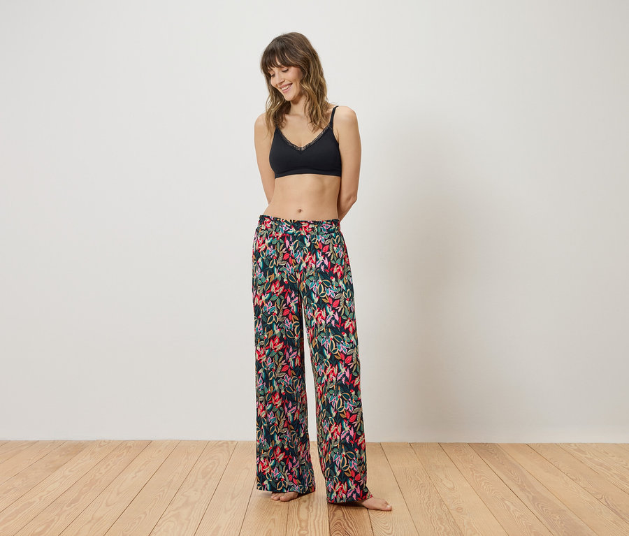 Siyah bralet ve çiçekli pantolonlu bir kadın beyaz bir duvarın önünde duruyor.