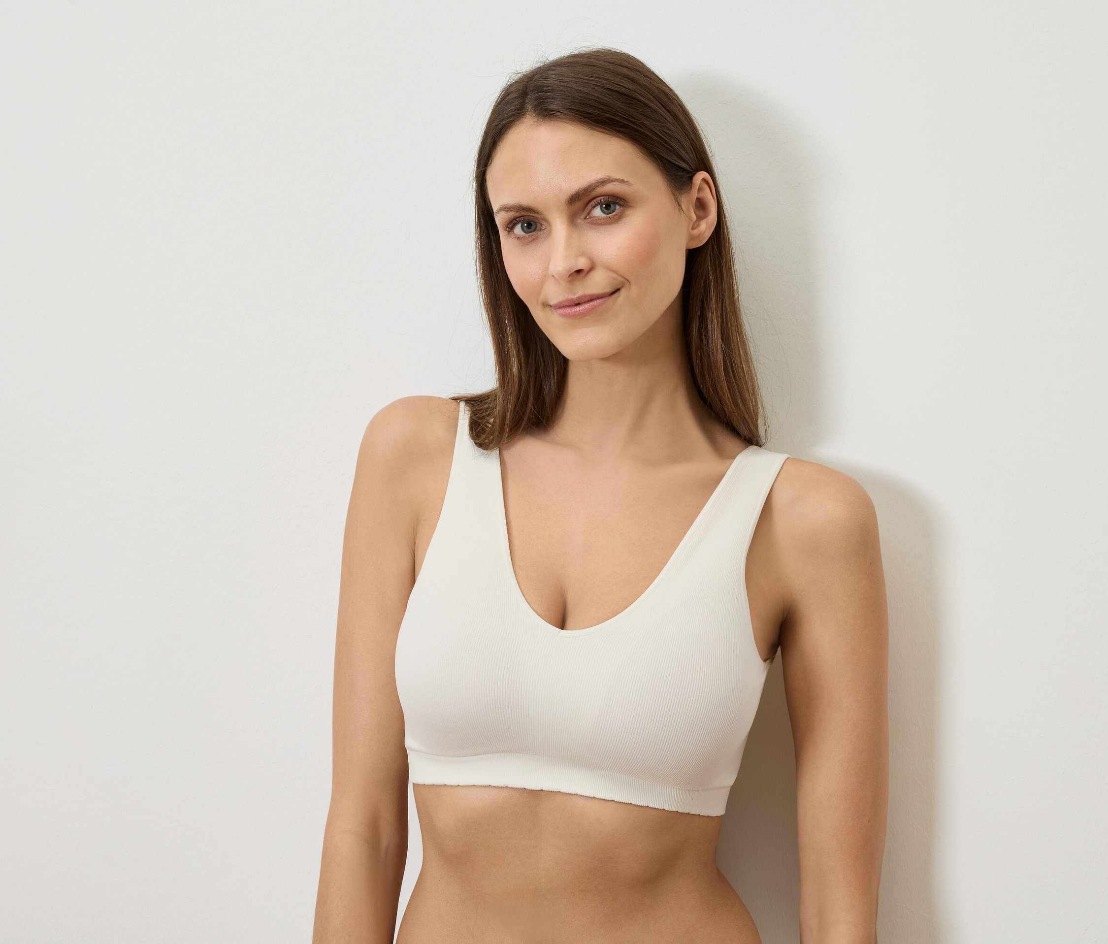 Uzun kahverengi saçlı bir kadın beyaz bir sütyen giymiş, beyaz bir duvara yaslanıyor.