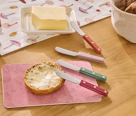Ahşap bir masanın üzerinde yumurtalıkta bir yumurta, tereyağı, tereyağlı bir rulo ve kırmızı, pembe, yeşil ve kırmızı renklerde dört tereyağı bıçağı duruyor.