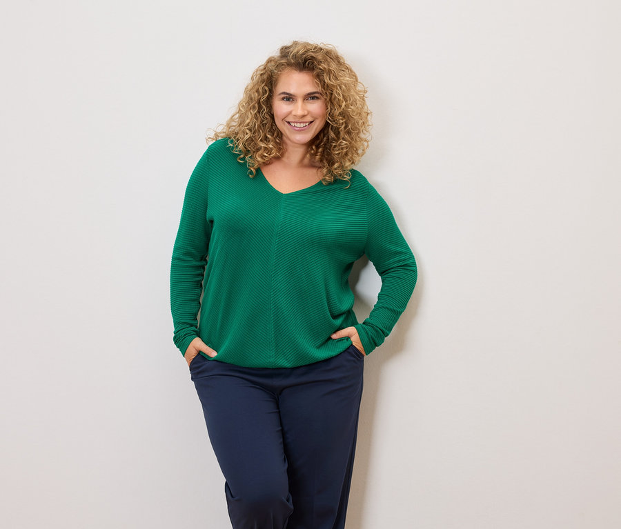 Kıvırcık saçlı bir kadın yeşil bir kazak ve lacivert pantolon giyiyor, elleri cebinde.