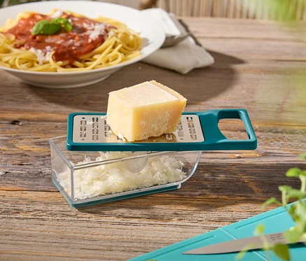 Ahşap bir masanın üzerinde bir tabak spagetti, peynirli bir peynir rendesi, fesleğenli ve kabaklı bir kesme tahtası duruyor.