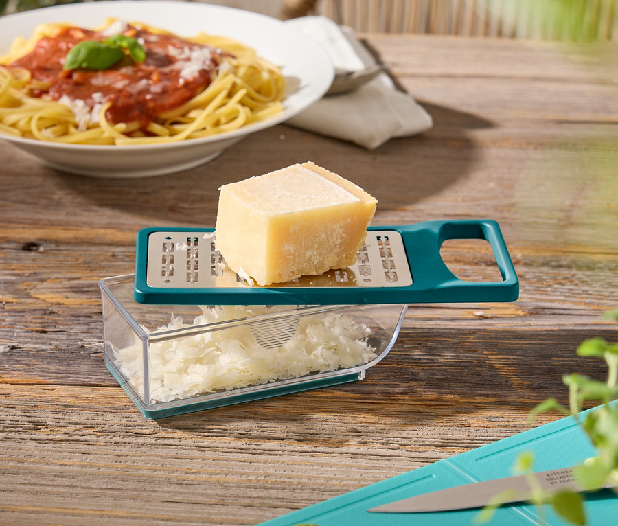 Ahşap bir masanın üzerinde bir tabak spagetti, peynirli bir peynir rendesi, fesleğenli ve kabaklı bir kesme tahtası duruyor.