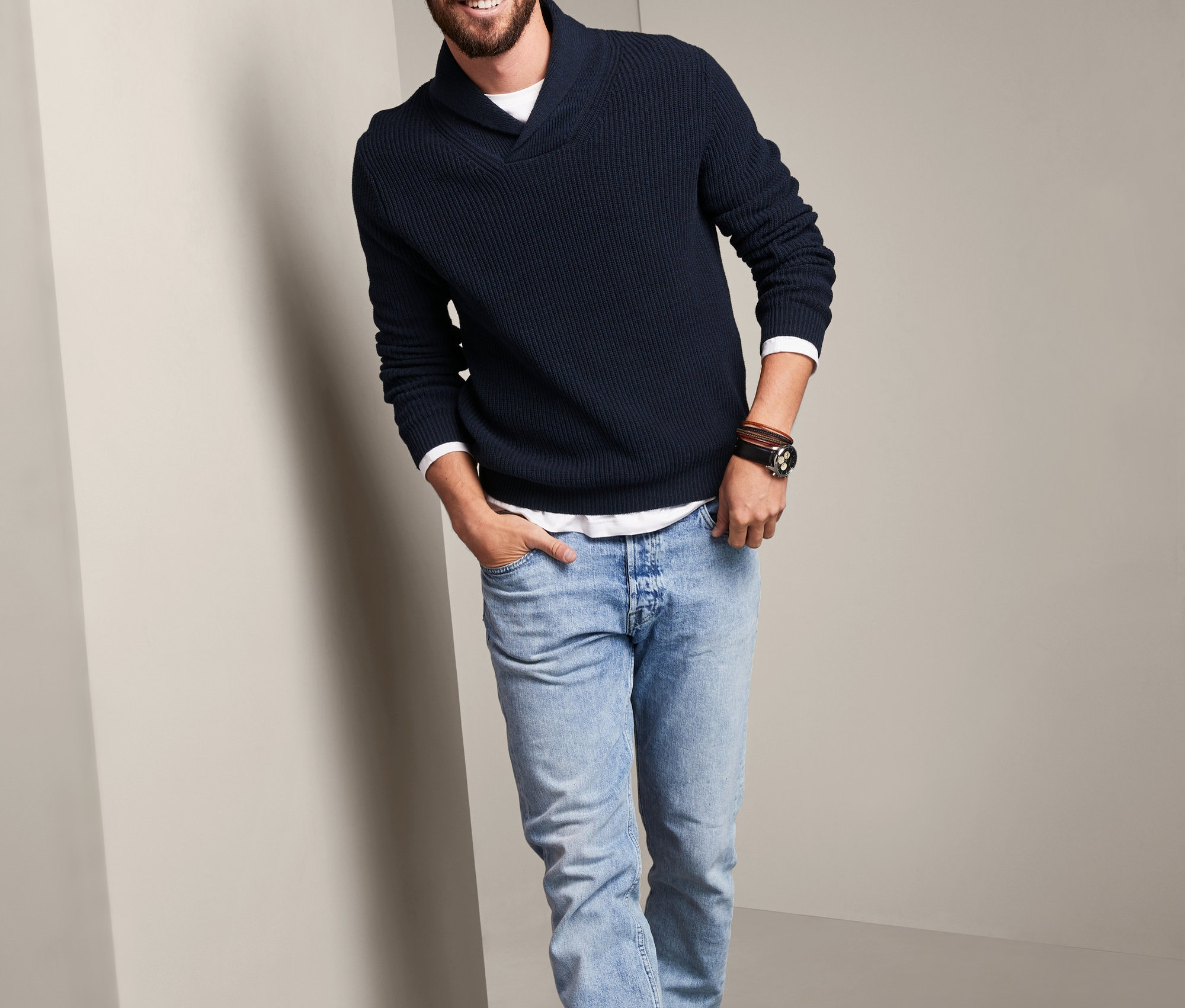 Sırıtan bir adam Organik Pamuklu Şal Yakalı İri Örgü Kazak, kot pantolon ve botlarla poz veriyor.