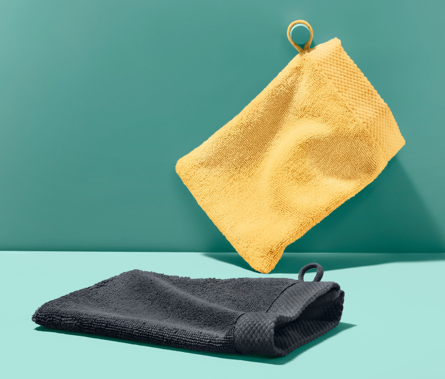 İki adet banyo lifi: sarı renkli olan asılı duruyor, antrasit renkli olan turkuaz yüzeyde duruyor.