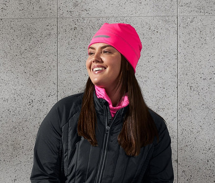 Gülümseyen bir kadın bankta oturuyor, üzerinde siyah termal ceket, termal koşu taytı, reflektör baskılı neon pembe termal bere var. Elinde bir su şişesi tutuyor.