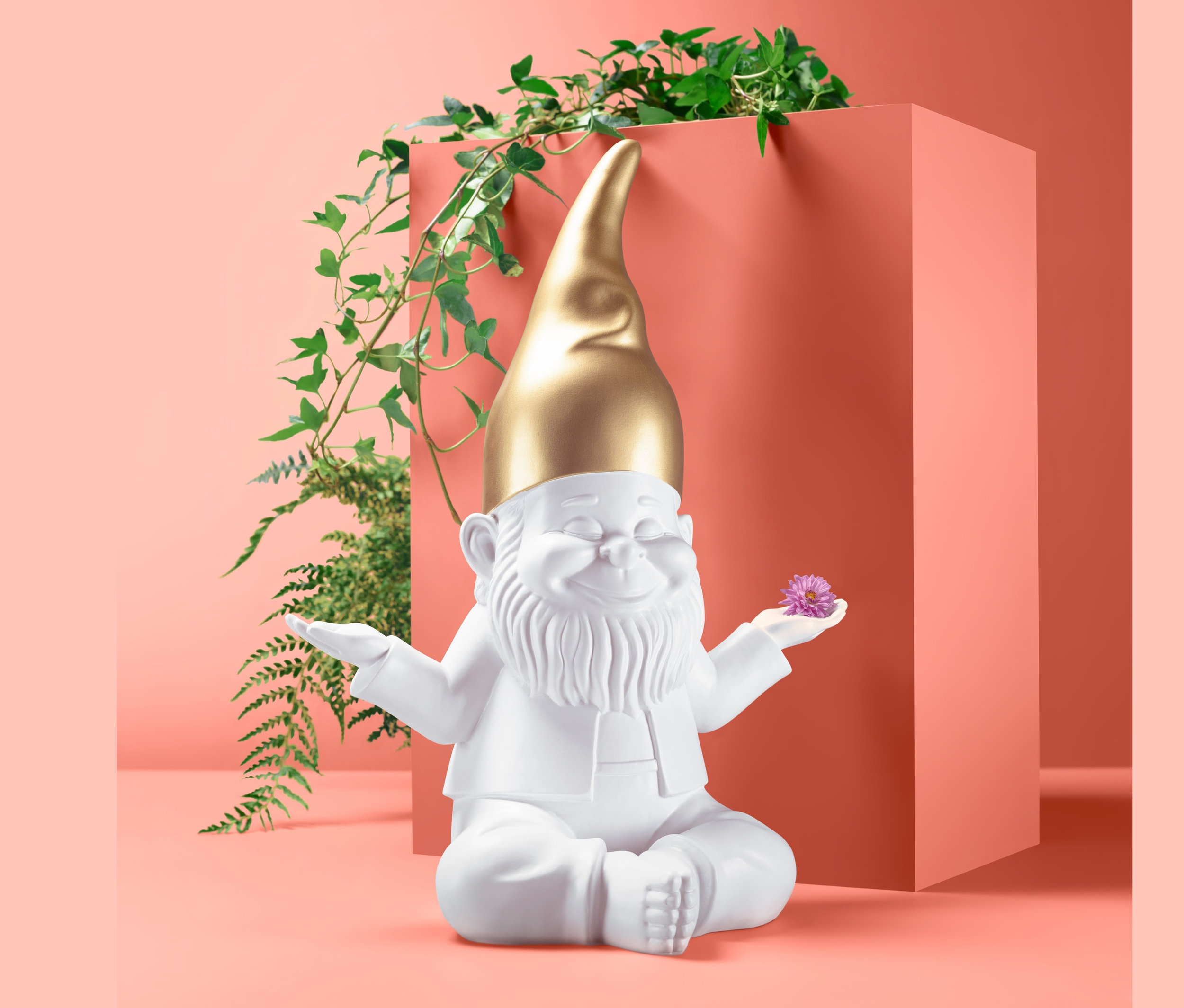 Altın şapkalı beyaz XXL dekoratif bahçe cücesi yerde oturuyor ve mor bir çiçek tutuyor.