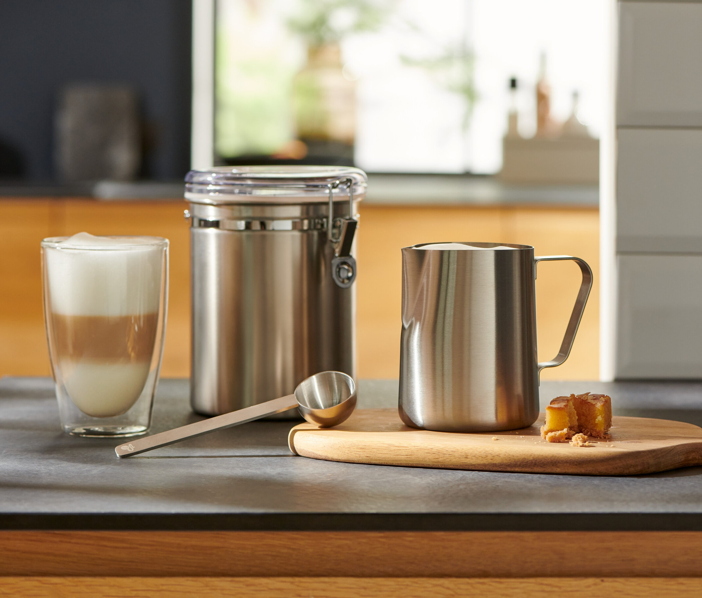 Mutfak tezgahında bir bardak sütlü kahve, gümüş bir kahve ölçü kaşığı, bir kap ve bir sütlük bulunmaktadır