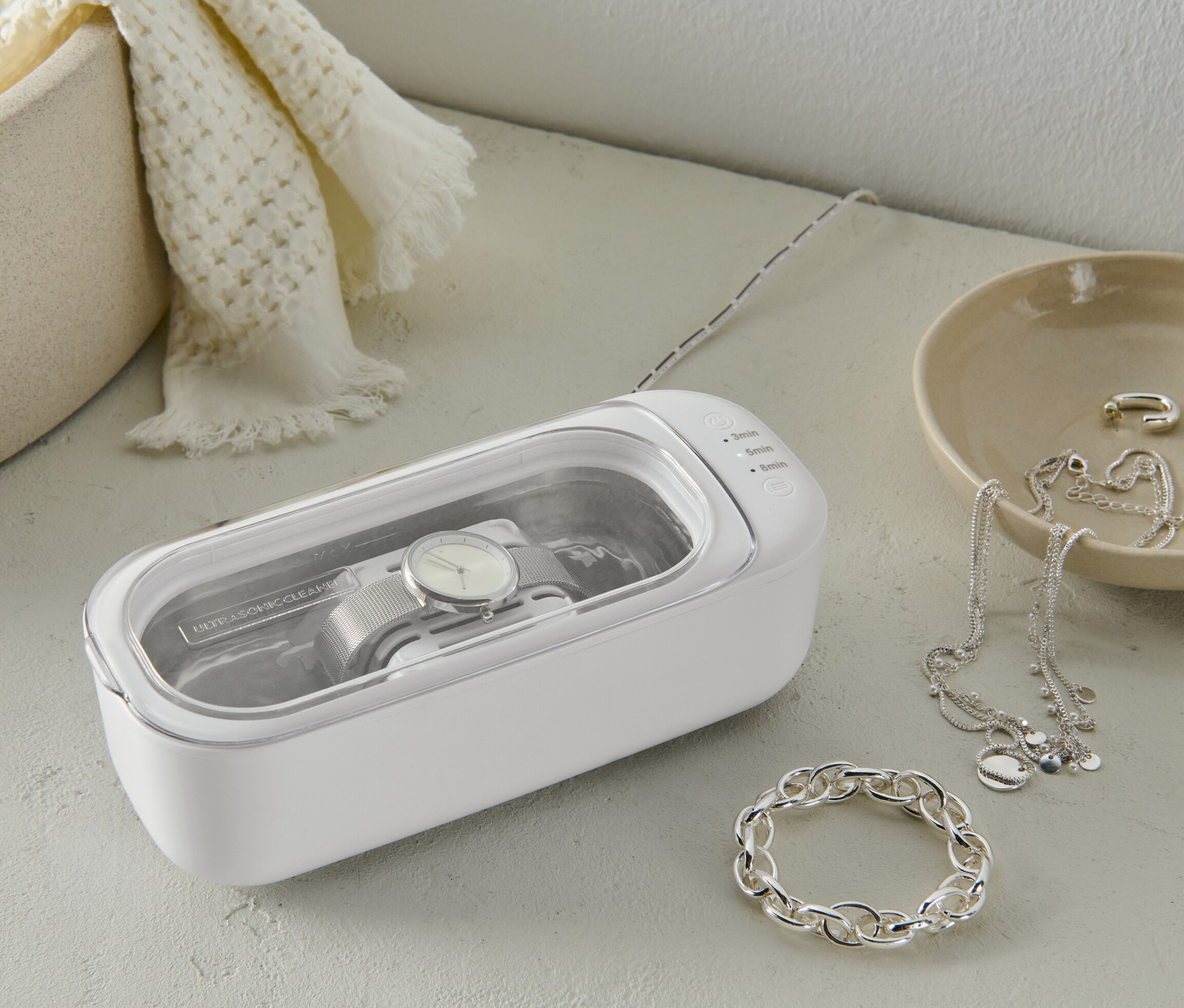 Beyaz bir ultrasonik temizleyicide temizlenen bir saat. Yanında mücevherler var.
