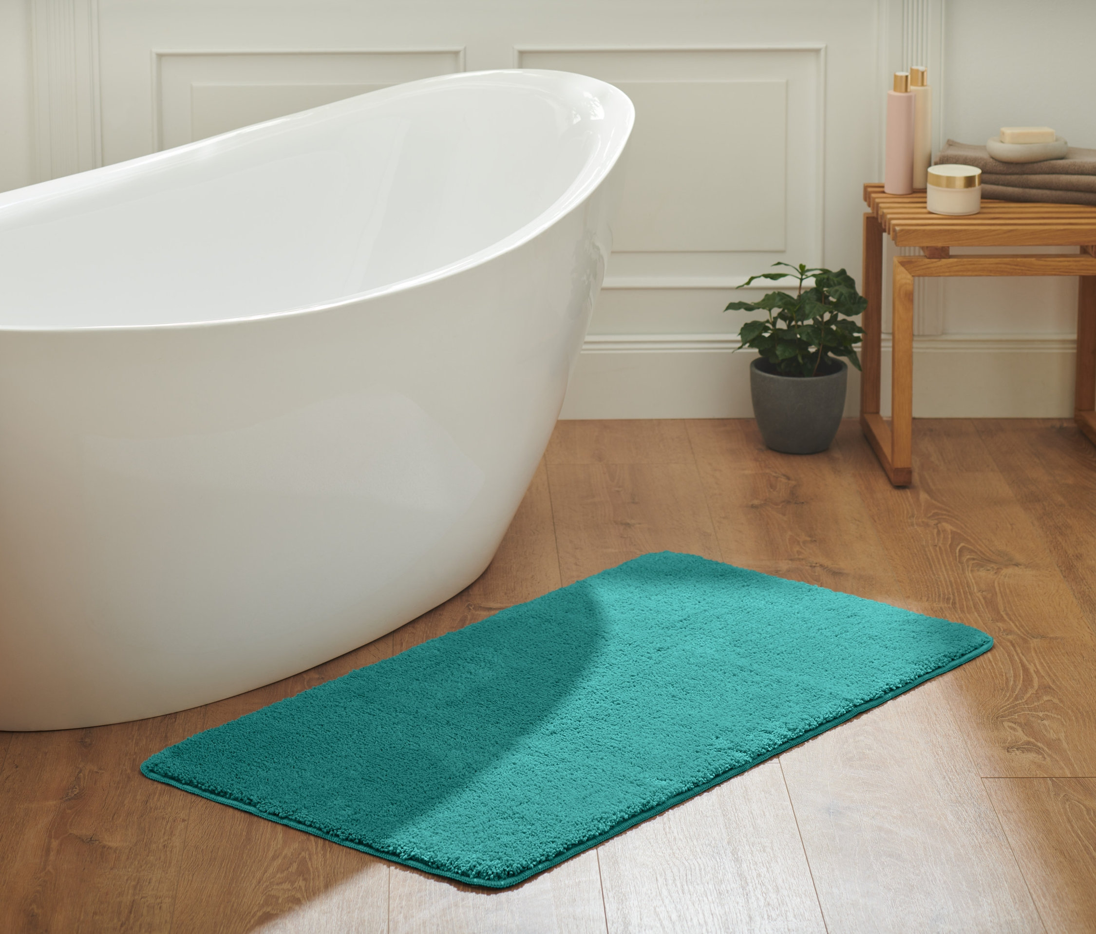 Ahşap zeminde havlu ve turkuaz banyo paspası olan ayaklı küvet.