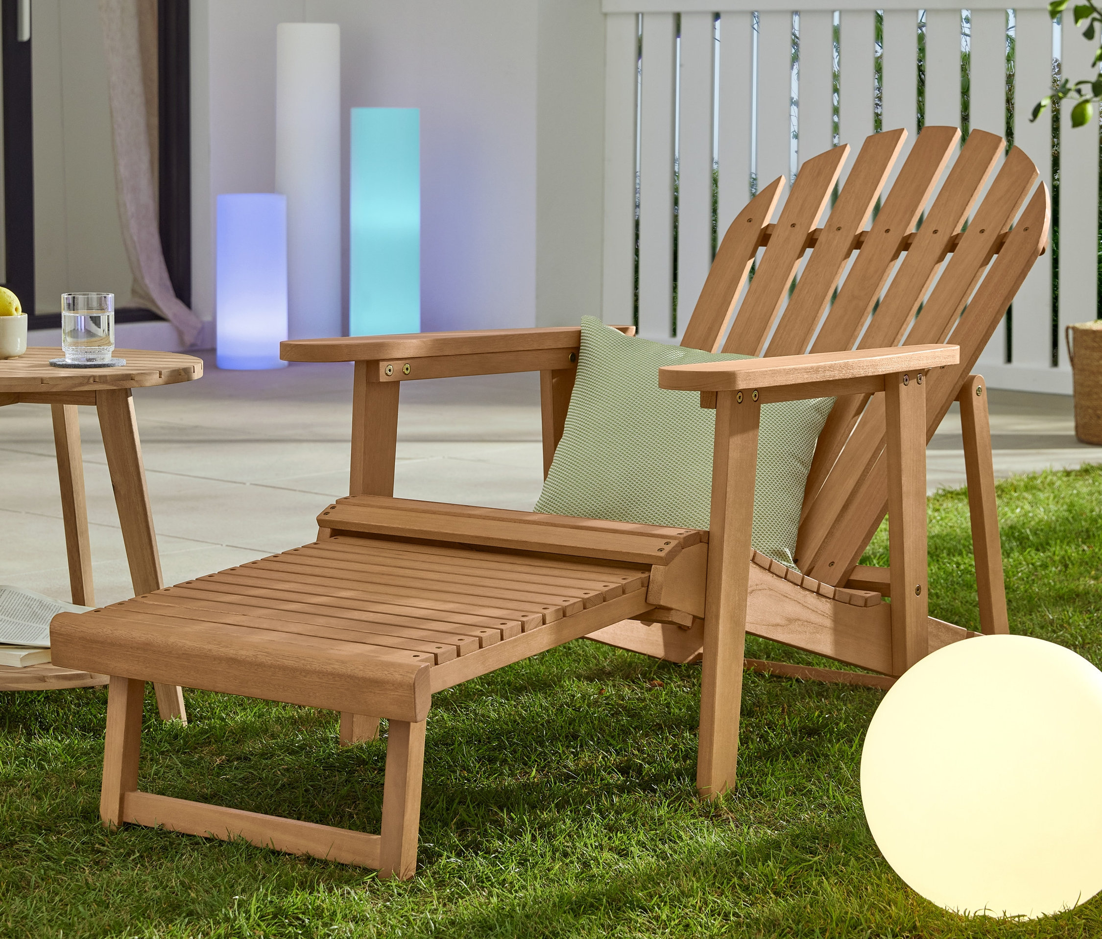 Çimde doğal renkli, uzatılabilir ayaklıklı bir Adirondack bahçe sandalyesi, tik görünümlü 2 kademeli bir »Lenja« yan sehpa ve solar küre lamba (yakl. 30 cm) bulunmaktadır.