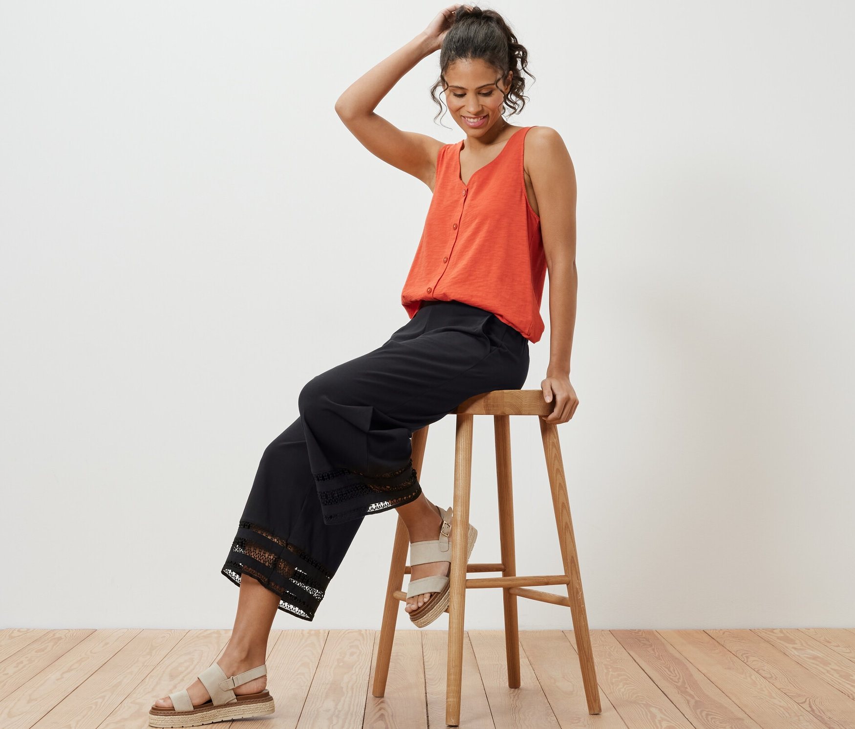 Kadın ahşap bir taburede oturuyor, turuncu bir atlet ve nakışlı siyah culotte pantolon giyiyor.