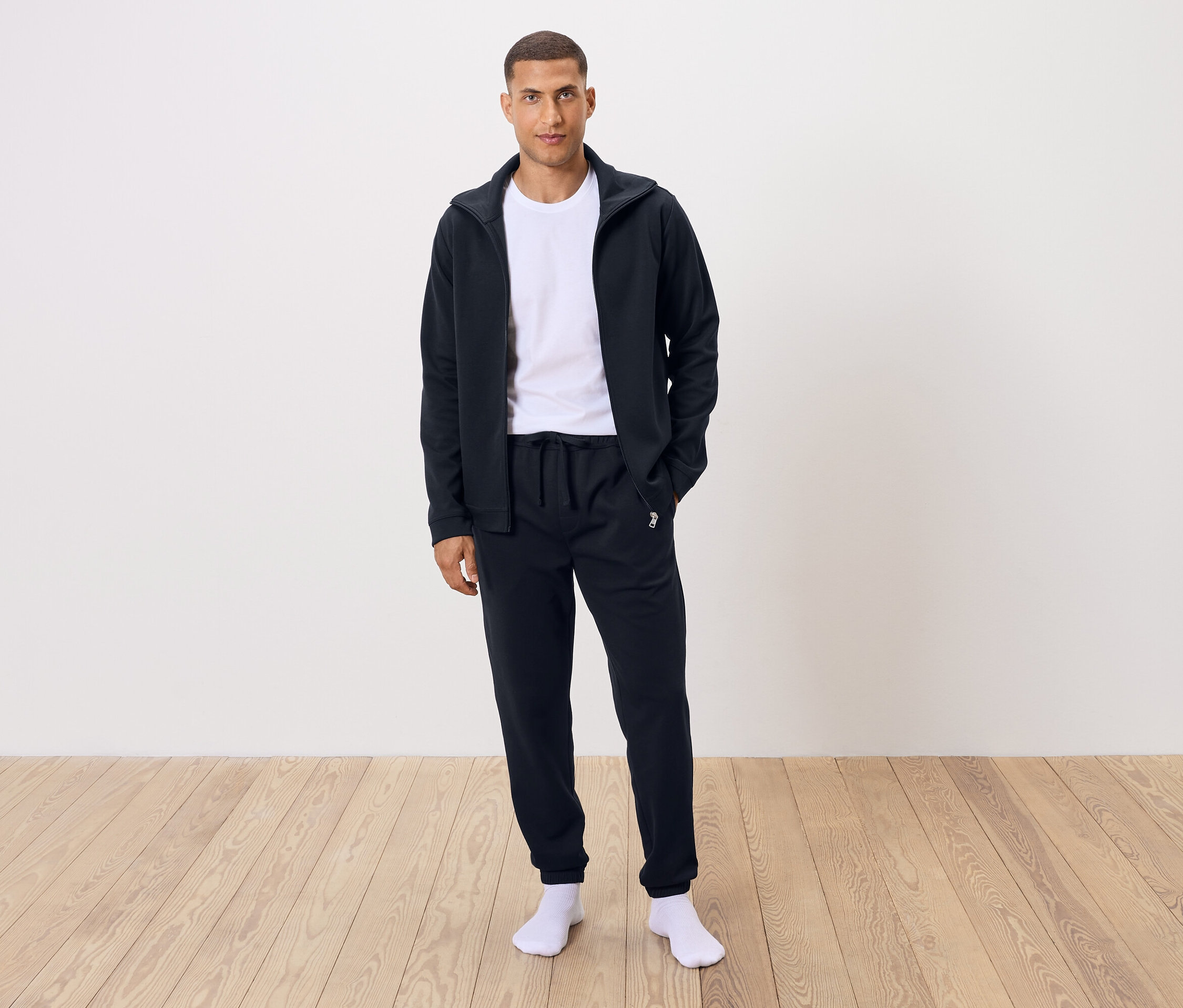 Beyaz tişört, siyah eşofman altı ve siyah fermuarlı ceket giyen bir adam ayakta duruyor.