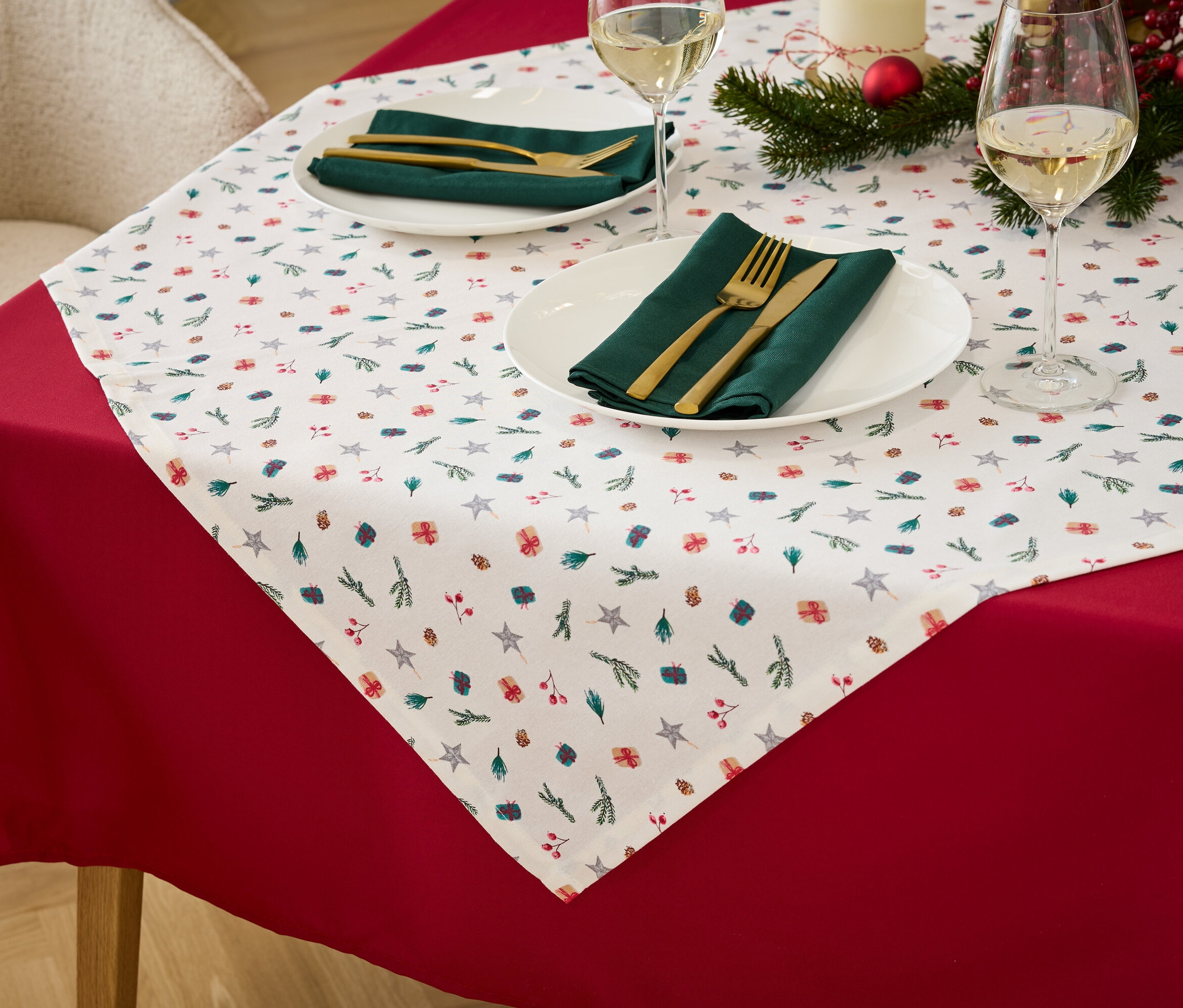 Kırmızı masa örtüsü, Noel desenli beyaz masa örtüsü, yeşil kumaş peçeteler ve altın rengi çatal bıçak takımı olan bir masanın detayı.