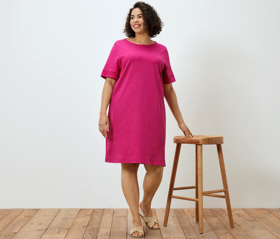 Bir kadın ahşap taburenin yanında poz veriyor ve pembe jarse elbise giyiyor.